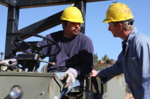 Two construction workers wearing yellow hard hats talking to each other while one is on a forklift.