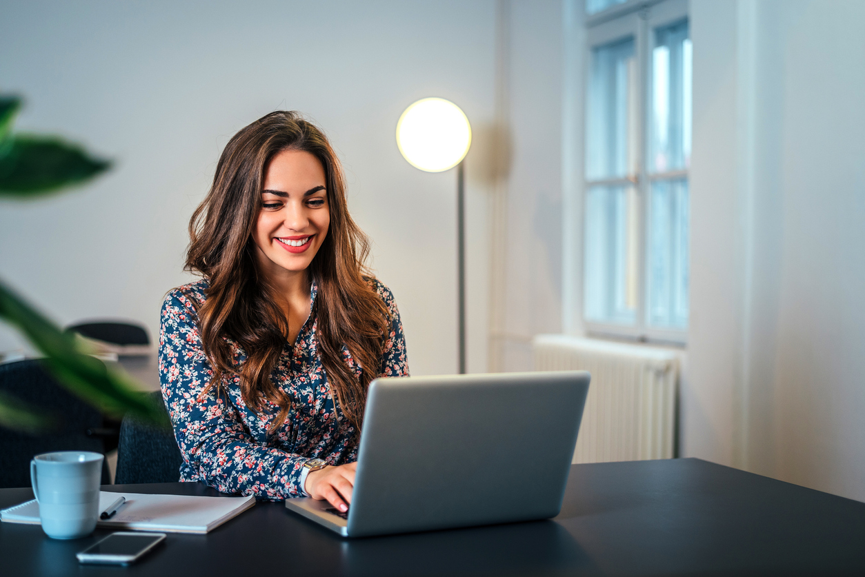 Young woman wearing flowered blouse, sitting at desk and smiling while typing on a silver laptop. Lamp and a window in the background.